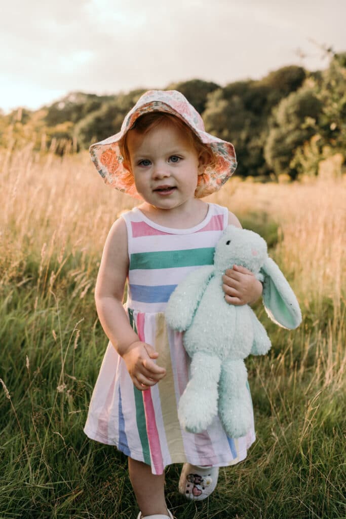 Little girl is walking in the field of long grass. She is wearing lovely stripy dress, flower hat and is holding her bunny. Family photographer in Hampshire. Ewa Jones Photography