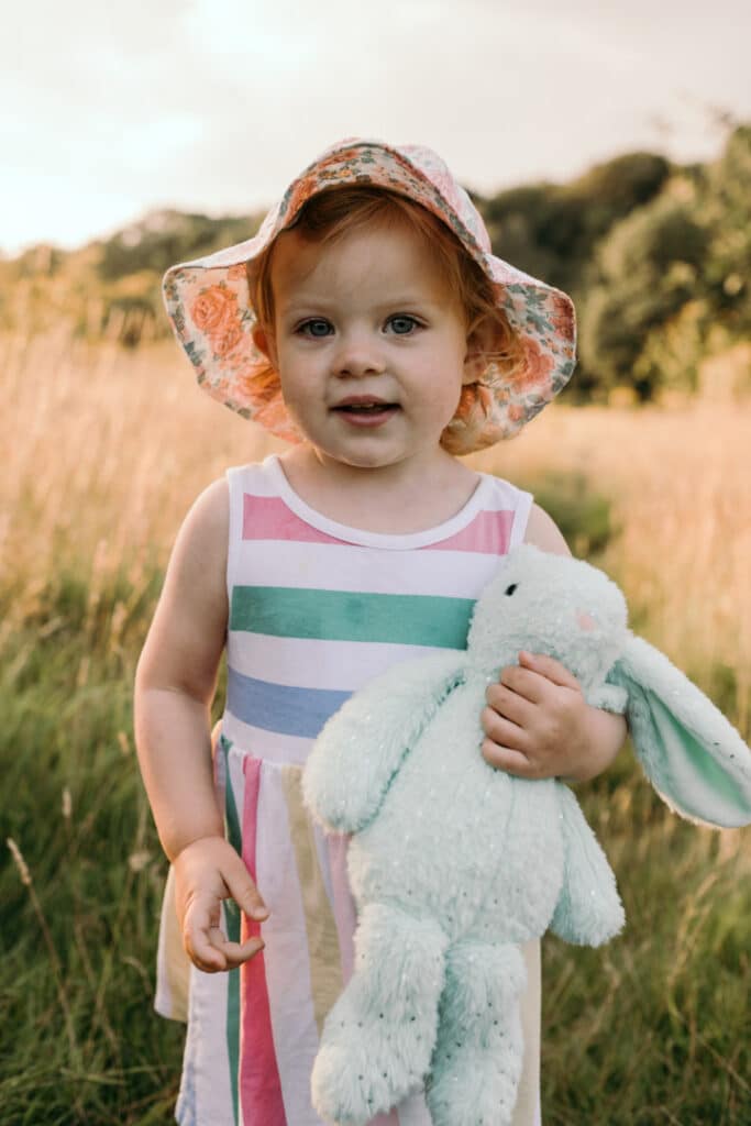 Little girl is walking in the field of long grass. She is wearing lovely stripy dress, flower hat and is holding her bunny. Family photographer in Hampshire. Ewa Jones Photography