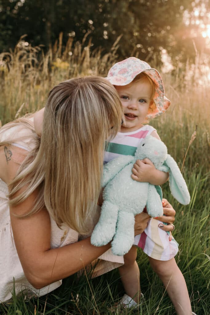 Mum is kissing her girl in her cheek. The sun is shining and making the gold sunset. Little girl is walking in the field of long grass. She is wearing lovely stripy dress, flower hat and is holding her bunny. Family photographer in Hampshire. Ewa Jones Photography