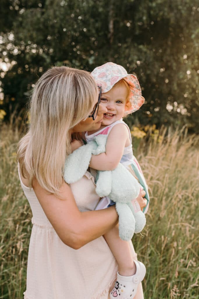 Cuddles with mummy. Little girl is cuddling to her mum and smiling to the camera. She is holding her rabbit toy and wearing stripy dress and flower hat. Family photography in Hampshire. Ewa Jones Photography