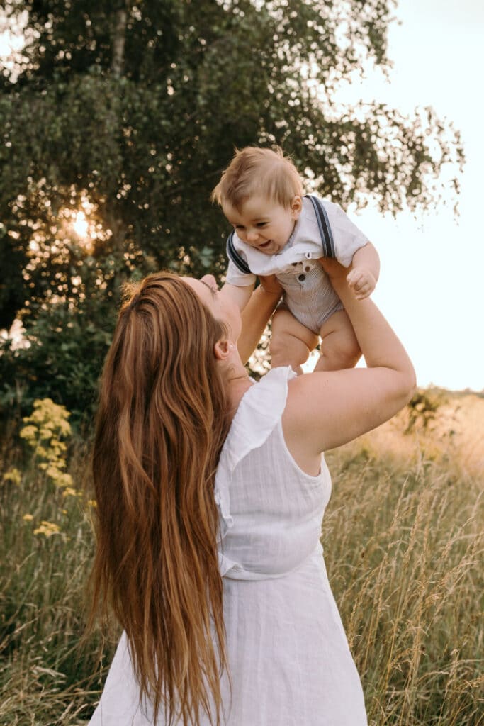 Mum is holding up her baby boy and looking at him. Baby boy is smiling back at his mum. Family photographer in Hampshire. Ewa Jones Photography