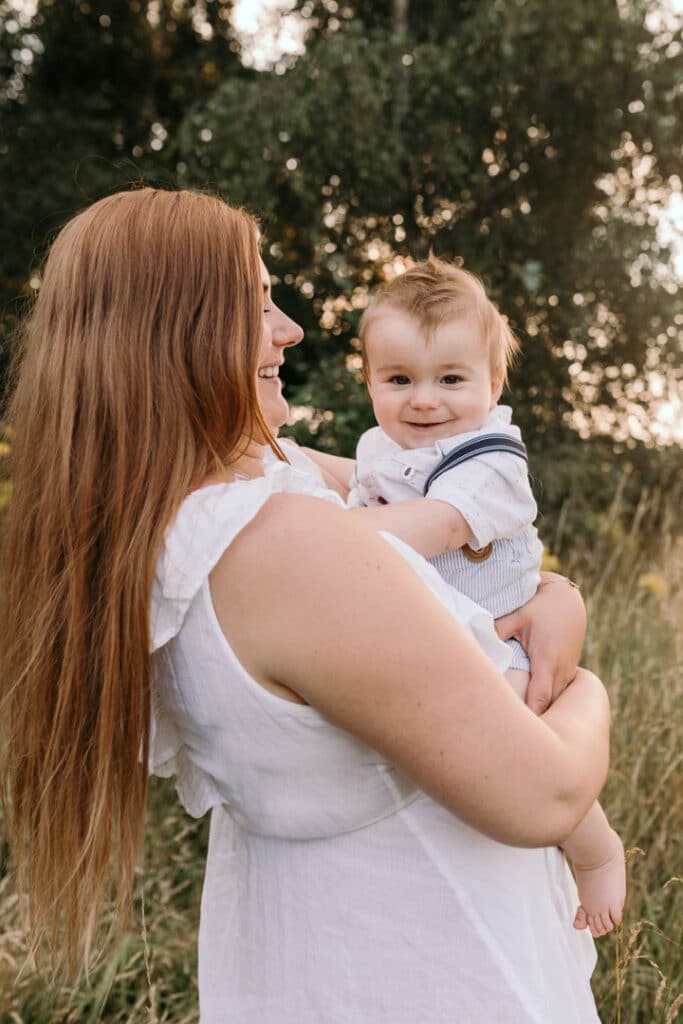 Mum is holding her little baby boy and looking at him. Boy is smiling at the camera. Lovely sunset family session. Family photographer in Hampshire. Ewa Jones Photography