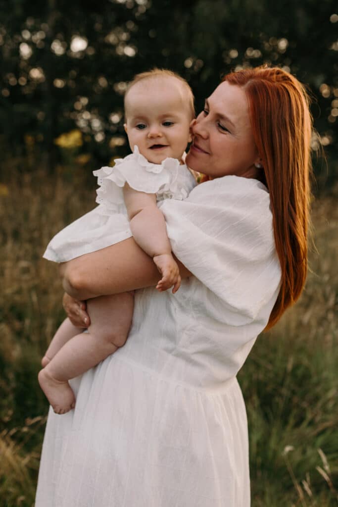 Mum is wearing lovely white dress and looking at her baby girl. Baby girl is also wearing lovely white top and smiling. Family photography in Basingstoke, Hampshire. Ewa Jones Photography