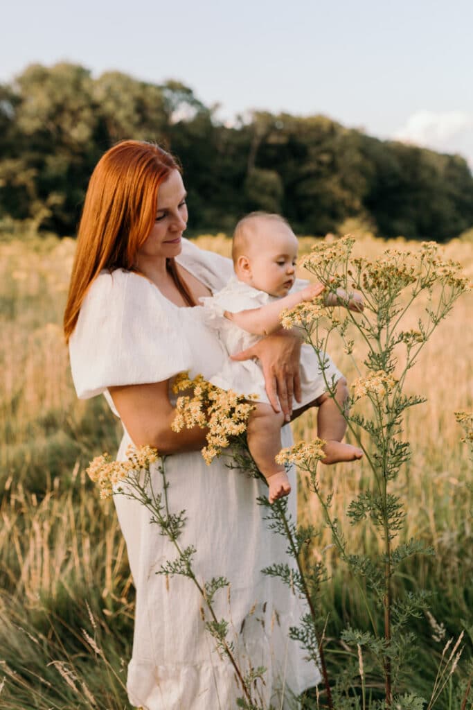 Mum is holding her baby girl and baby girl is touching wild flowers on the field. Lovely sun is shining on mmmy's back. Family photographer in Hampshire. Ewa Jones Photography