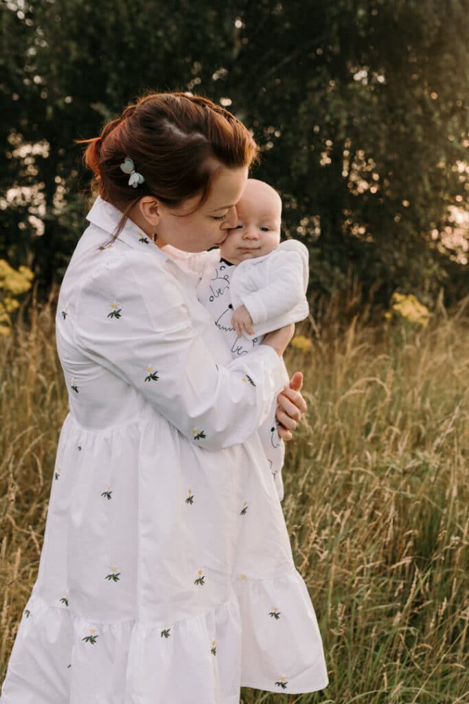 Mum is kissing her baby boy on his cheek. Mum is wearing lovely white dress with white daisies. Sunset photo shoot in the wild flower field. Family photography in Basingstoke, Hampshire. Ewa Jones Photography