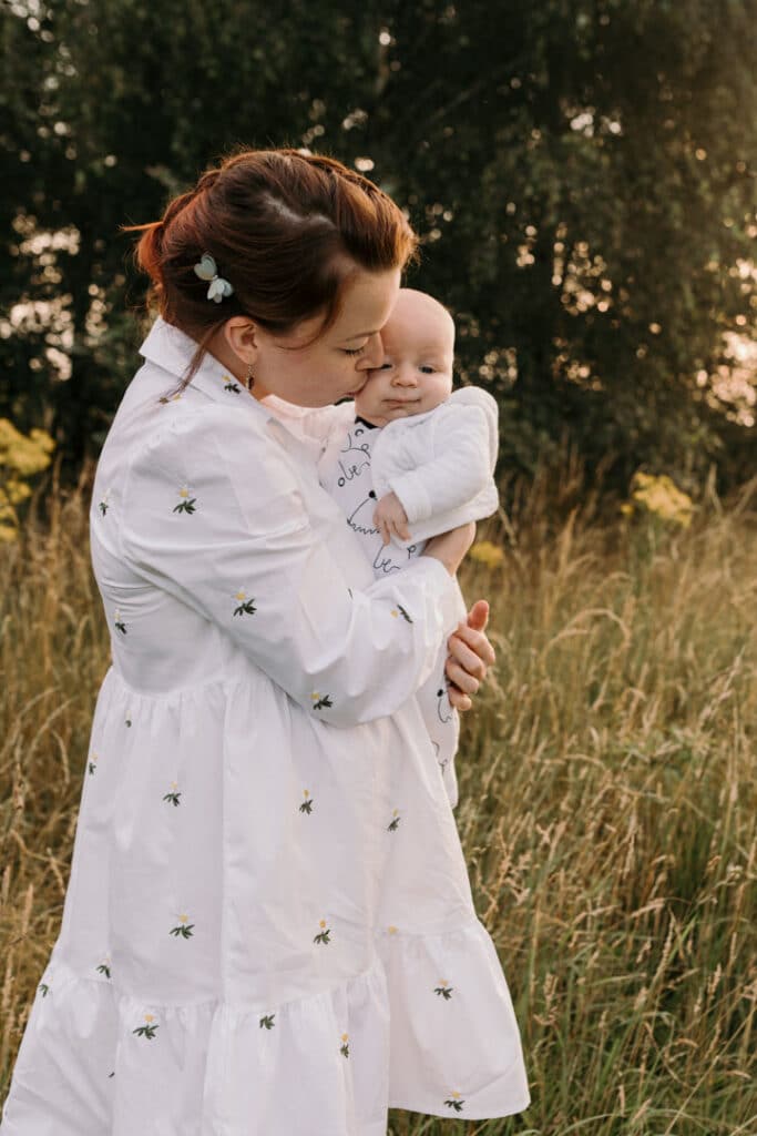 Mum is kissing her baby boy on his cheek. Mum is wearing lovely white dress with white daisies. Sunset photo shoot in the wild flower field. Family photography in Basingstoke, Hampshire. Ewa Jones Photography