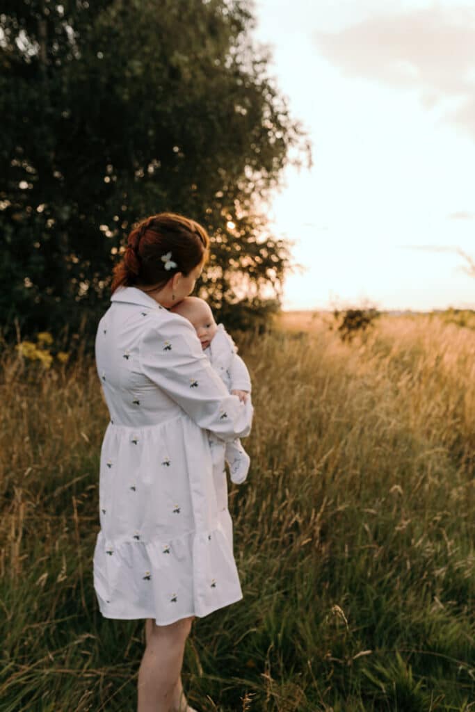 Mum is cuddling her baby boy and looking at the sunset. Mum is kissing her baby boy on his cheek. Mum is wearing lovely white dress with white daisies. Sunset photo shoot in the wild flower field. Family photography in Basingstoke, Hampshire. Ewa Jones Photography