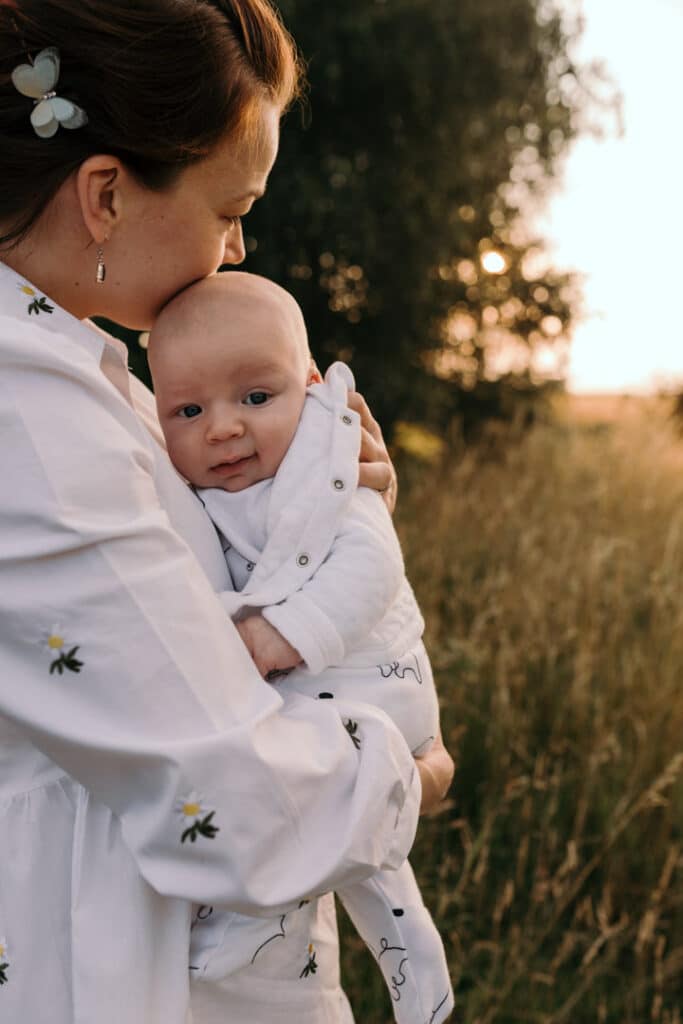 Mum is cuddling her baby boy and looking at the sunset. Mum is kissing her baby boy on his cheek. Mum is wearing lovely white dress with white daisies. Sunset photo shoot in the wild flower field. Family photography in Basingstoke, Hampshire. Ewa Jones Photography