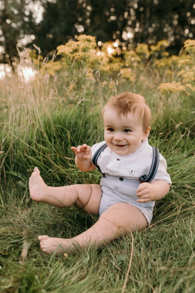 boy is sitting on the grass and smiling. he is looking at his mum and wearing lovely white t-shirt and stripy shorts. Family photo shoot in Basingstoke, Hampshire. Ewa Jones Photography