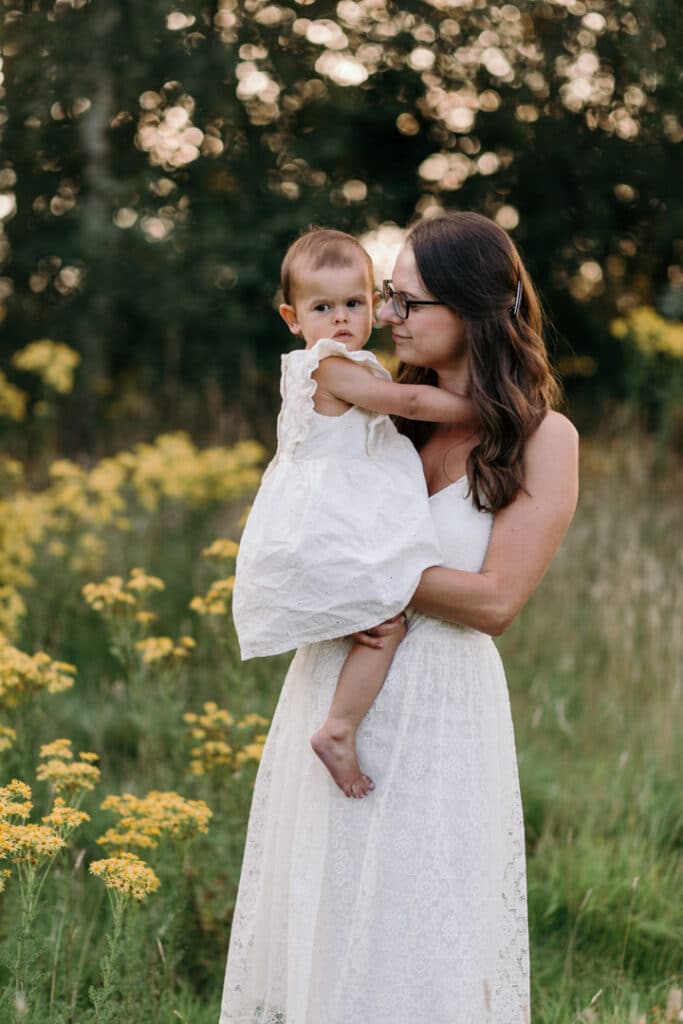 Mum is holding her little girl in her arms a looking at her. Sun is shining and mum is wearing white dress with blue flowers and girl is wearing lovely pink dress. Family photographer in Basingstoke, Hampshire. Ewa Jones Photography