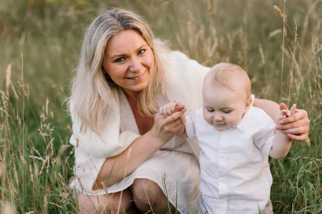 Mum is holding her little boy hands and mum is looking at the camera. She is wearing a lovely white dress and boy is wearing white t-shirt. Lovely sunset photo session. Family photographer in Hamsphire, Ewa Jones Photography