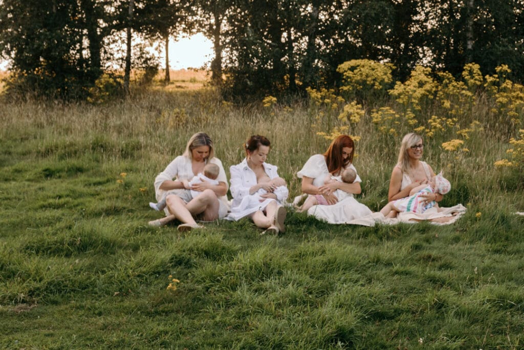 four mums are sitting on the blanket and breastfeeding their babies. There is a gorgeous field full of wild flowers and the sun is shining through the trees. Group breastfeeding photo session in Basingstoke, Hampshire. Ewa Jones Photography