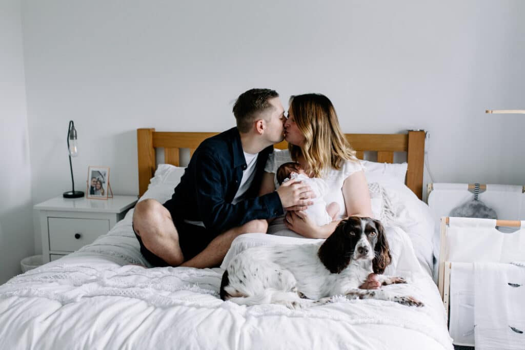 Mum and dad are sitting on the bed and giving each other a kiss. Mum is holding newborn baby girl and dog is looking at the camera. In home newborn photography in Hampshire. Ewa Jones Photography