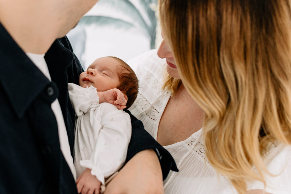 Dad is holding newborn baby girl and looking at her. Mum is standing next and also looking at her beautiful baby girl. Newborn photo session in Basingstoke, Hampshire. Ewa Jones Photography