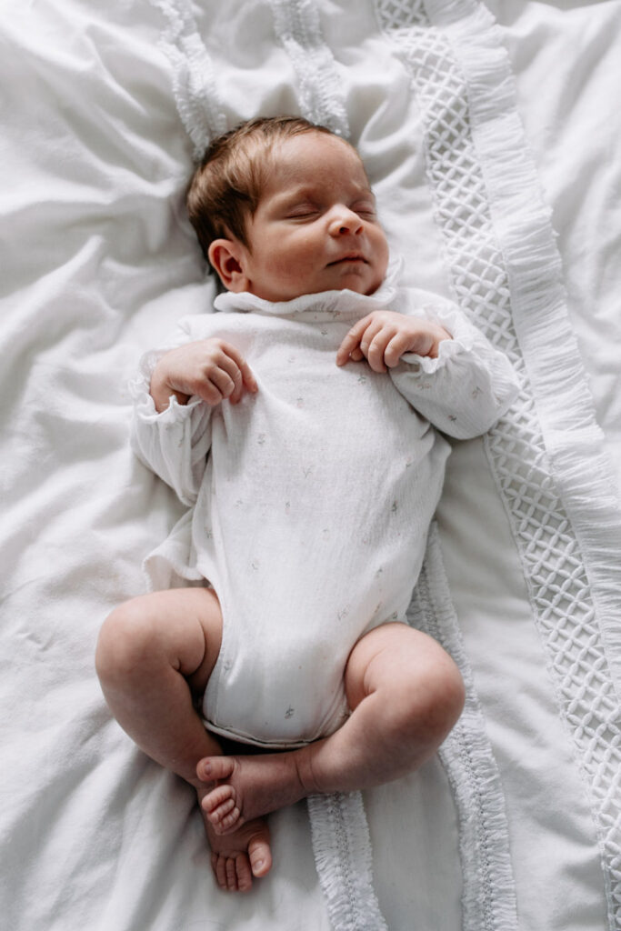 Newborn baby girl is sleeping on bed and smiling. She is wearing lovely white baby grow and sleeping on white bedding. Newborn photographer in Hampshire. Ewa Jones Photography