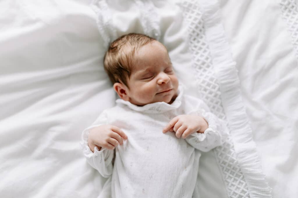 Newborn baby girl is sleeping on bed and smiling. She is wearing lovely white baby grow and sleeping on white bedding. Newborn photographer in Hampshire. Ewa Jones Photography
