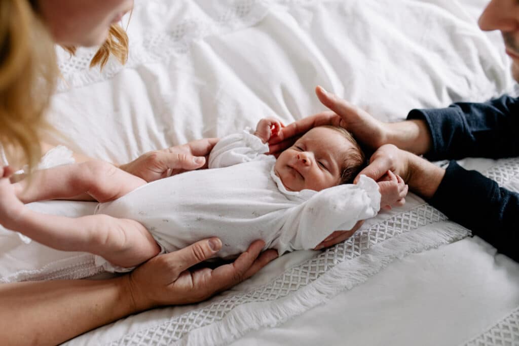 Newborn baby girl is laying on bed and both parents are holding baby girl from each side. In home newborn photography in Hampshire. Ewa Jones Photography