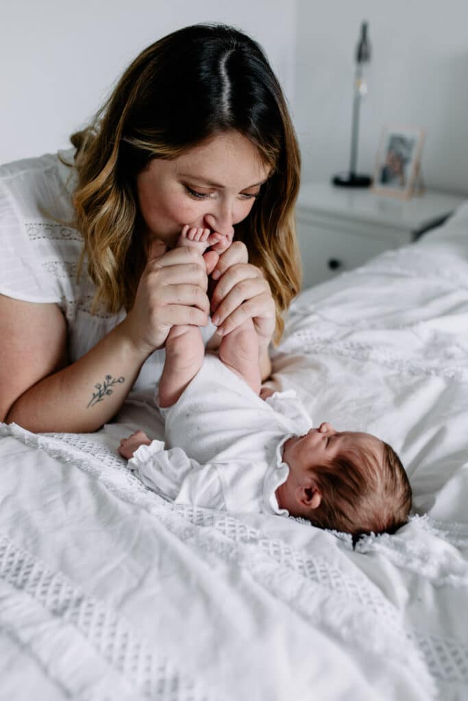 Mummy is kissing her newborn baby girl feet and looking at her. Newborn baby girl is laying on bed. Newborn photography in Basingstoke, Hampshire. Ewa Jones Photography