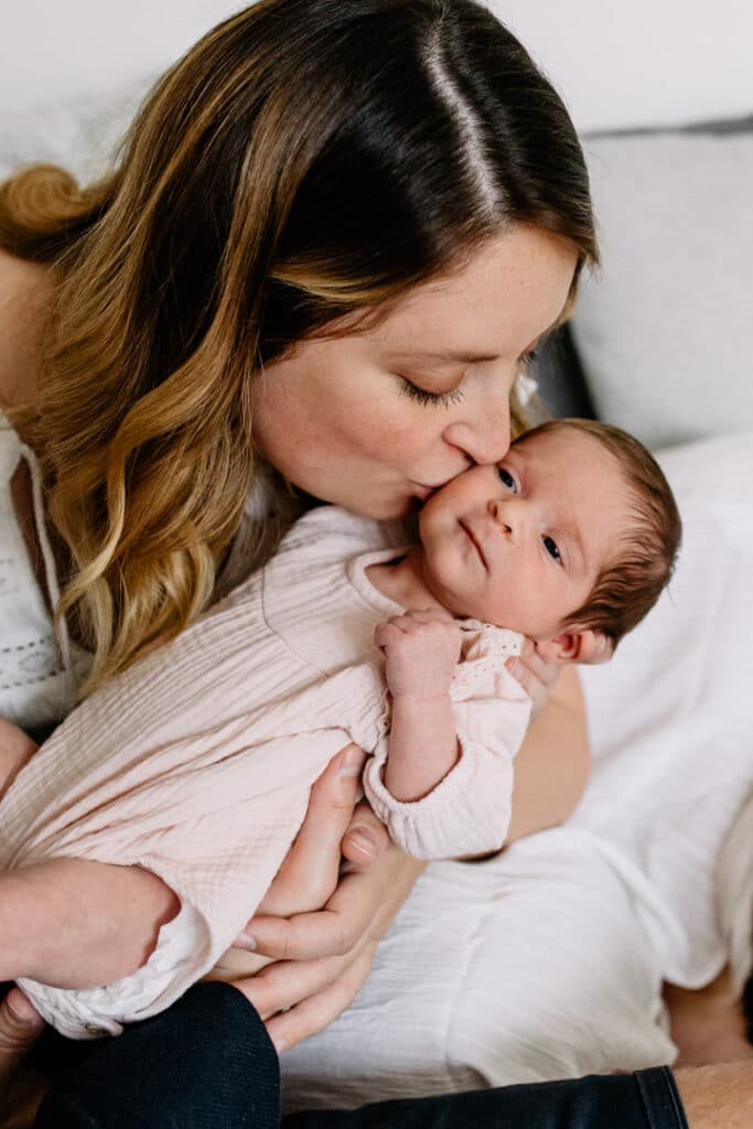 Mum is kissing her newborn baby girl on her cheek. Newborn baby is wearing lovely cream baby grow. Newborn photographer in Hampshire. Ewa Jones Photography