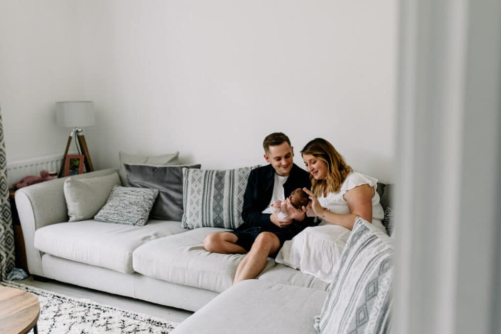 Mum and dad are sitting on the sofa and looking at their baby girl. Candid photograph of a new family. Natural in home lifestyle newborn photography in Hampshire. Ewa Jones Photography