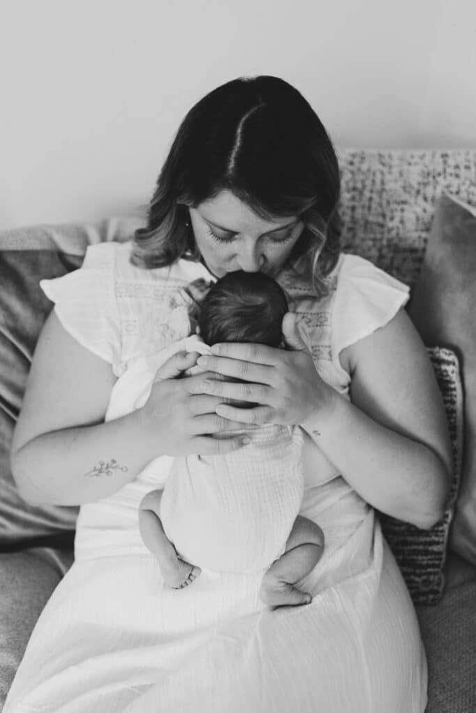 Mum is sitting on the sofa and kissing her newborn baby girl on top of her head. Newborn baby photography in Basingstoke, Hampshire. Ewa Jones Photography