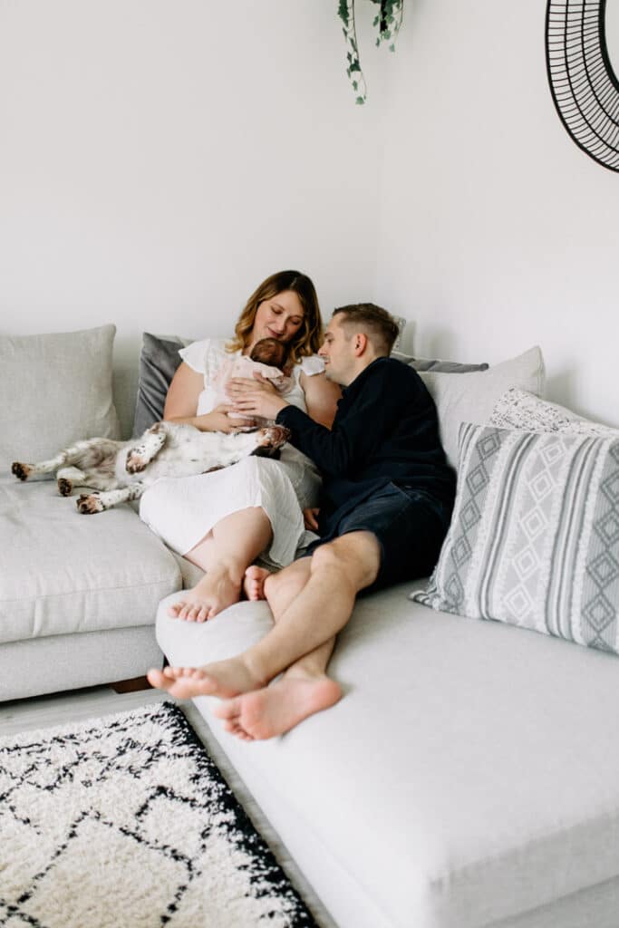 Mum and dad are both sitting on the sofa and looking at their newborn baby girl. Natural, family lifestyle photography in Basingstoke, Hampshire. Ewa Jones Photography