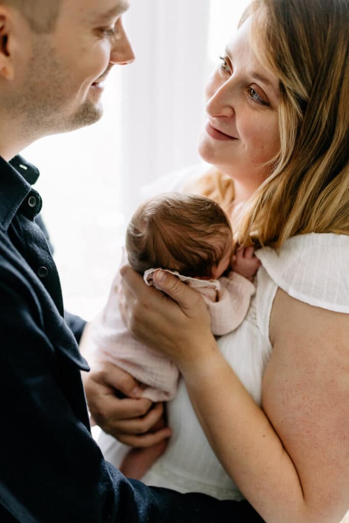 Mum and dad are standing next to the window and mum is holding her newborn baby girl. Mum is also looking lovingly at her husband. Lifestyle newborn photography in Hampshire. Ewa Jones Photography