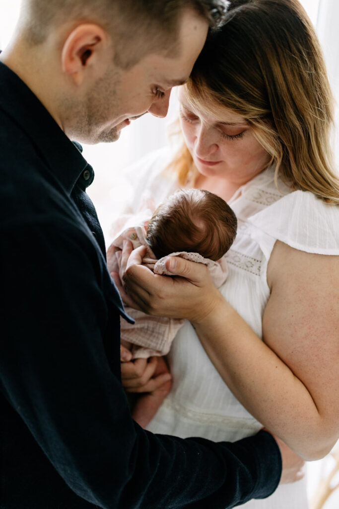 Mum and dad are close to each other and touching foreheads. Mum is holding her newborn baby girl in her arms. Newborn photographer in Basingstoke, Hampshire. Ewa Jones Photography