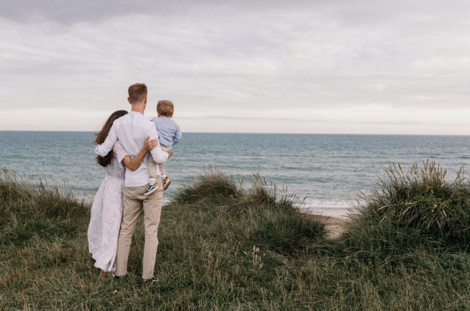 family of three is looking at the sea and holding each other. Lovely summer evening by the beach in Hampshire. family photo session at the beach. Ewa Jones Photography