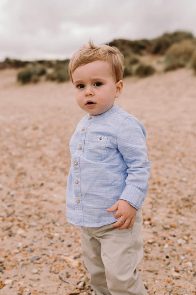 Little boy is standing on the sand and looking at the camera. He is wearing lovely blue shirt and cream trousers. Family photo sessions at the beach in Hampshire. Hampshire family photographer. Ewa Jones Photography