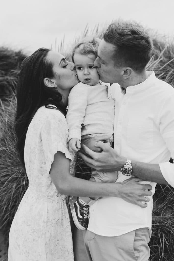 mum and dad are at the beach and dad is holding his son. Both parents are kissing little boy on his cheeks. Lovely candid family photo session in Hampshire by the beach. Ewa Jones Photography