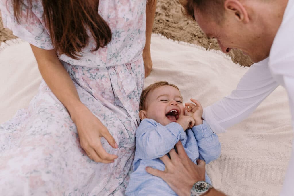 little boy is laying on the blanket and laughing as his dad is tickling him. Natural and unposed family photography in Hampshire. Ewa Jones Photography