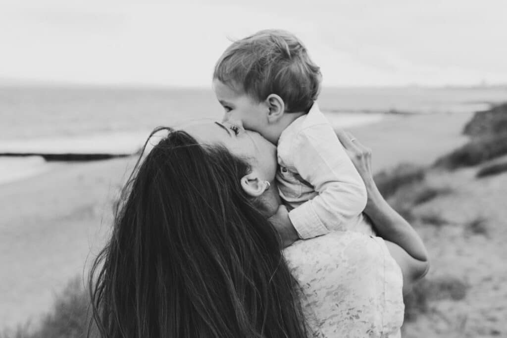 Mum is kissing her little boy on his cheeks. Lovely black and white image of mother and son love. Family photo shoot in Hampshire by the beach. Ewa Jones Photography