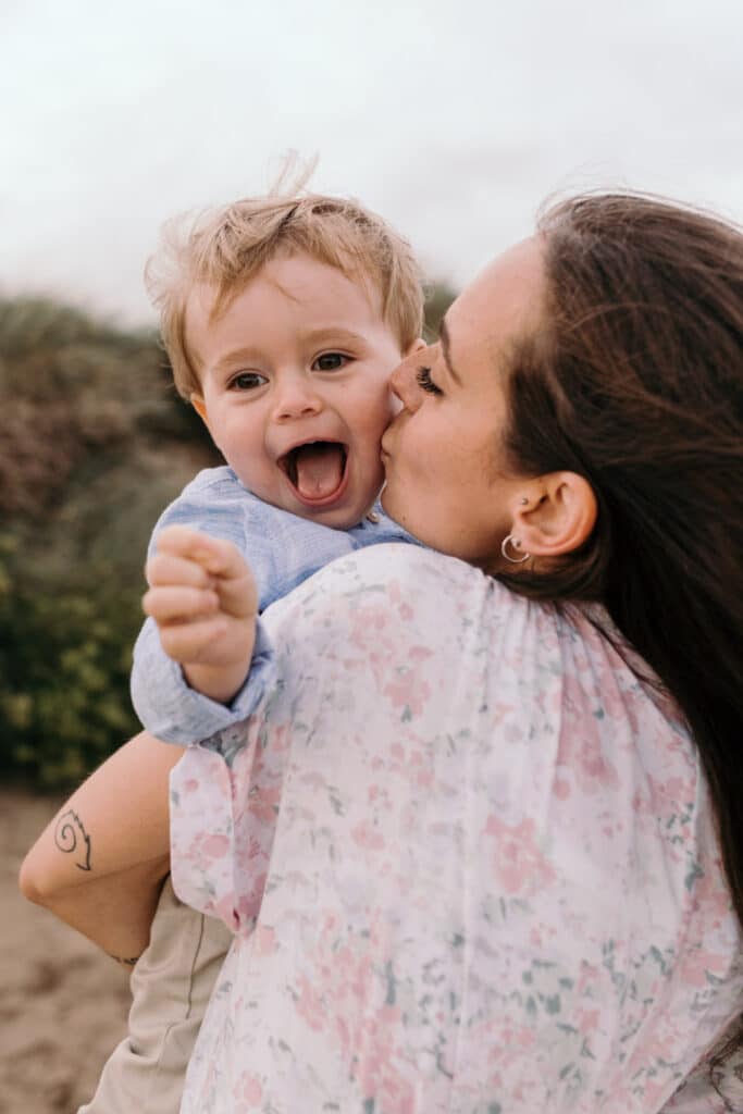 Little boy is smiling to the camera and mum is kissing his cheek. family photo session at the beach. Ewa Jones Photography