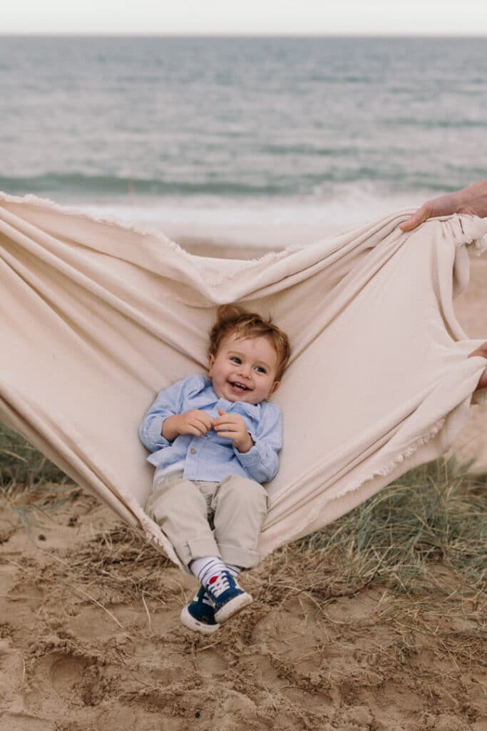 Little boy is laying on the blanket and mum and dad are swinging him. He is smiling and giggling. photo session is at the beach in Hampshire. family photo session at the beach. Ewa Jones Photography