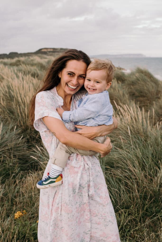 Mum is holding her little boy and laughing with him. They are standing on the sand dunes by the Hampshire beach. Family photo shoot in Hampshire. Ewa Jones Photography