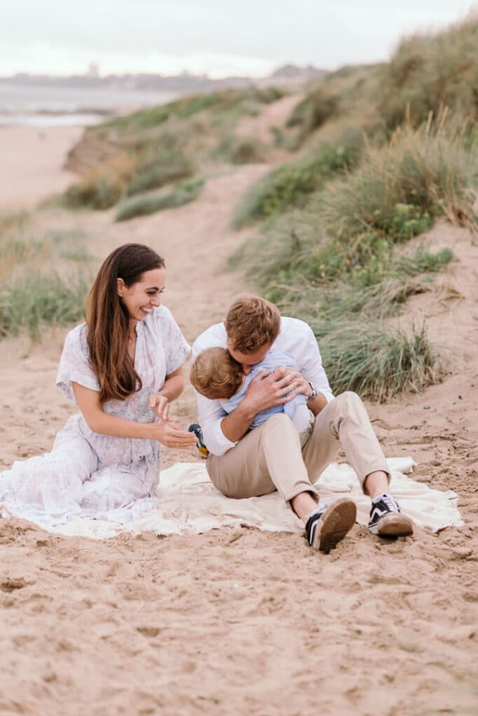 mum and dad are sitting on the blanket and dad is kissing his little boy and tickling him. Natural family photography in Hampshire. family photo session at the beach. Ewa Jones Photography