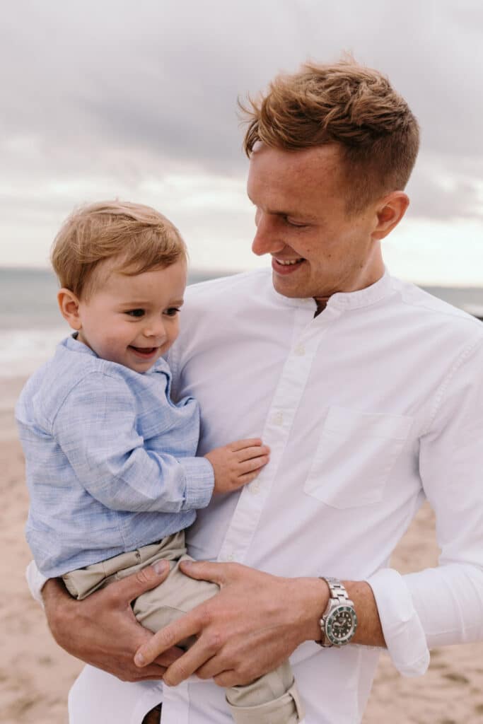 Dad is holding his son, looking at him and smiling to him. the little boy is smiling too. Family photo session at the beach in Hampshire. Ewa Jones Photography