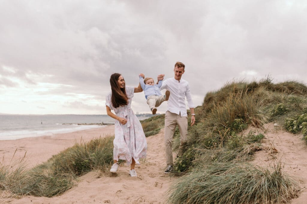 Mum and dad are swinging their little boy in the air. they are at the beach and around sand dunes. Family photography in Hampshire. Ewa Jones Photography