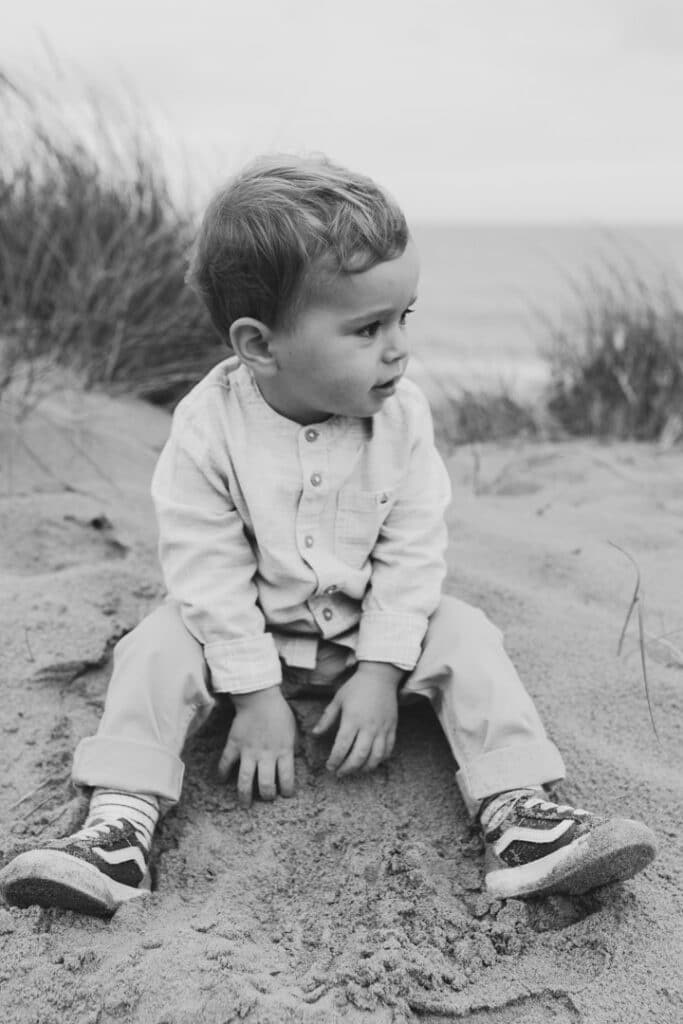 Little boy is sitting on the sand and touching the sand. He is looking away. Lovely black and white photograph. Family photographer in Hampshire. Ewa Jones Photography