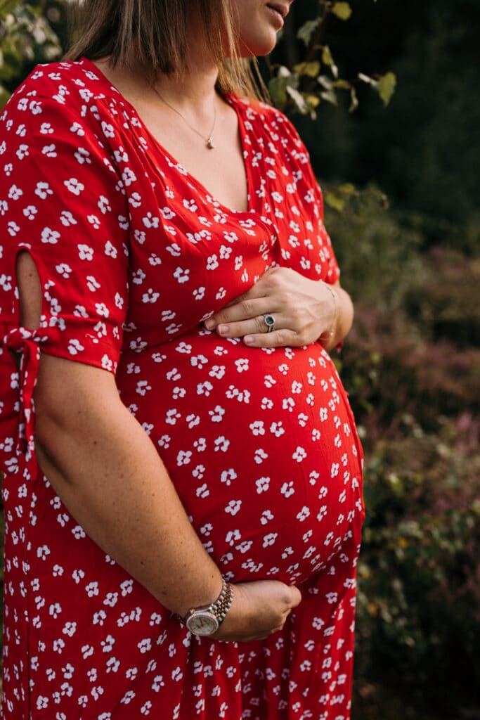 Expecting mum is holding her baby bump. she is wearing lovely maternity dress in red with white flowers. Maternity photo session in Hampshire. Ewa Jones Photography
