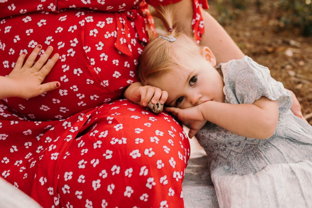 Little girl is resting her head on mummy's leg. she is looking at the camera. Her older sister is touching baby bump. maternity photo shoot in Hampshire. Ewa Jones Photography