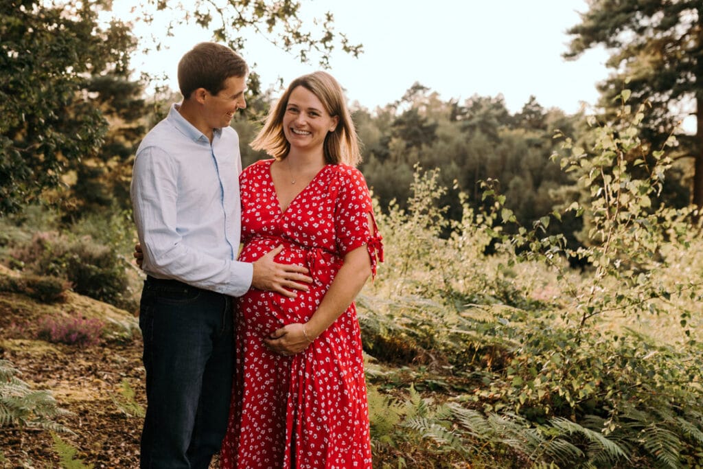 Maternity photo session in Hampshire. Mum is holding her baby bump and looking at the camera. she is smiling and husband is looking at her and smiling. lovely candid moment. Maternity photo session in Hampshire. Ewa Jones Photography