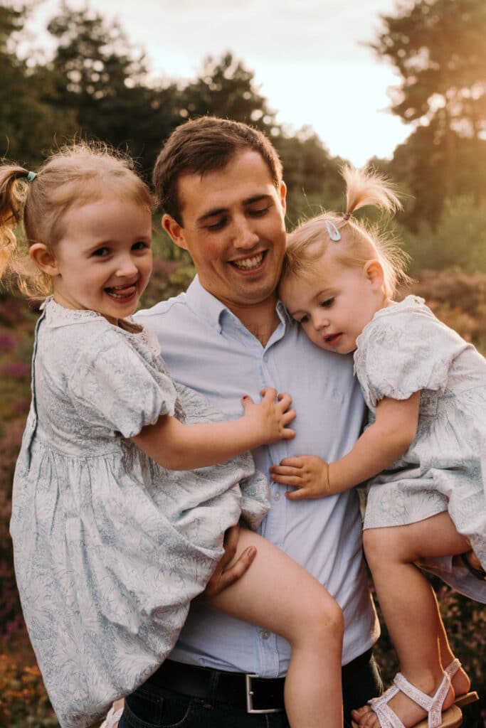 Dad is holding his two little girls on his arms. One daughter is giggling and another is cuddling up to daddy. Lovely candid family photo session in Hampshire. Ewa Jones Photography