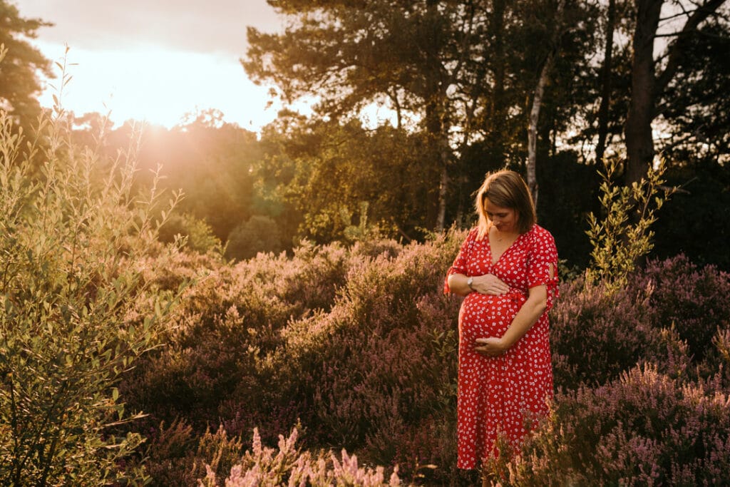 Expecting mum is looking down at her baby bump and holding her bump. It's a lovely summer evening and sun is shining behind mum. Maternity photoshoot in Hampshire. Ewa Jones Photography