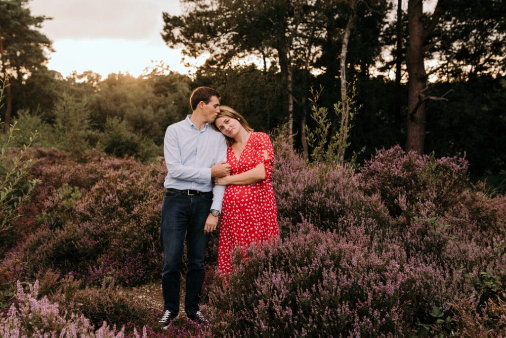 expecting parents are standing in the fields of lovely purple flowers. Pregnant mum is wearing a red dress with white flowers. dad is kissing mum on top of her head. maternity photo session in Hampshire. Ewa Jones Photography