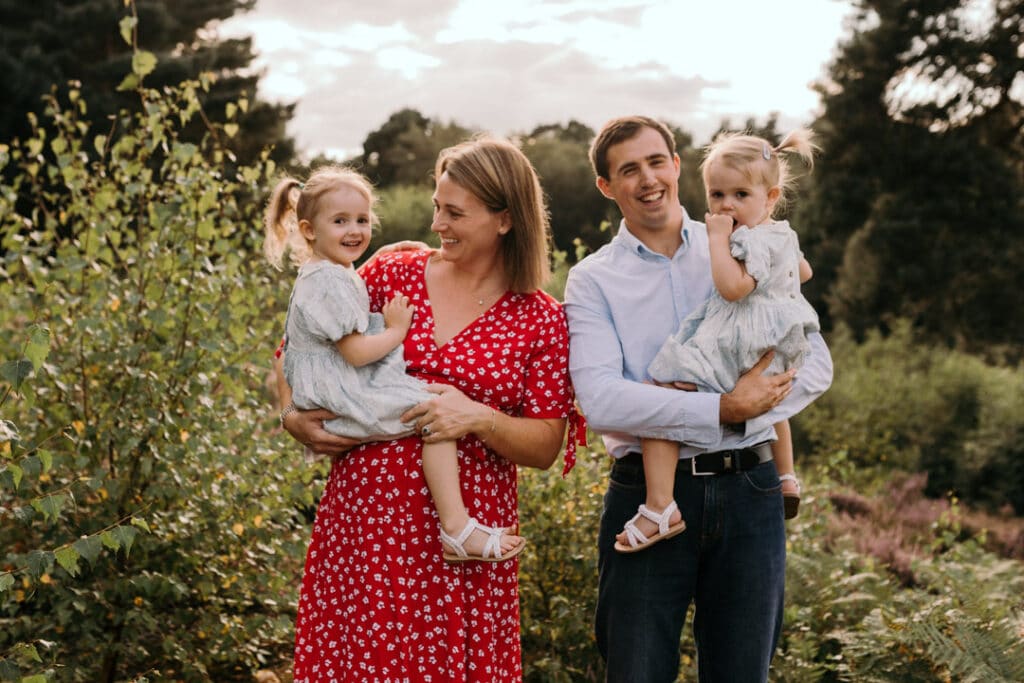 family of four is standing and smiling. Mum is holding older daughter and dad is holding younger daughter. lovely family photo session in Hampshire. Ewa Jones Photography
