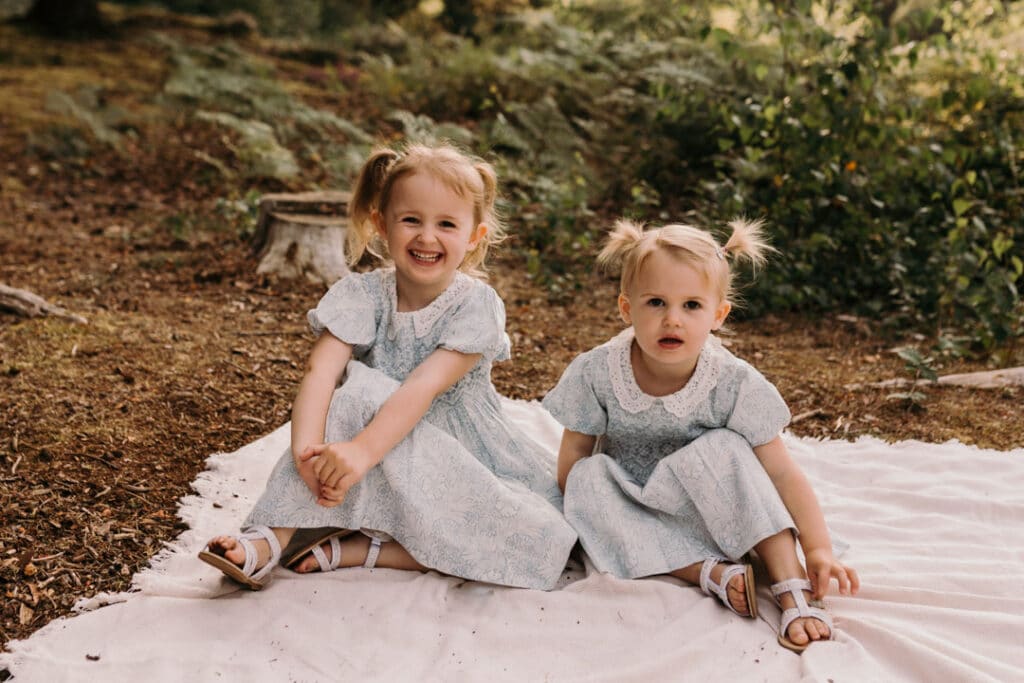 two sisters are sitting on the blanket and looking at the camera. they bothe are wearing the same blue and white dresses and two ponytails. lovely family photography in Hook, Hampshire. Ewa Jones Photography