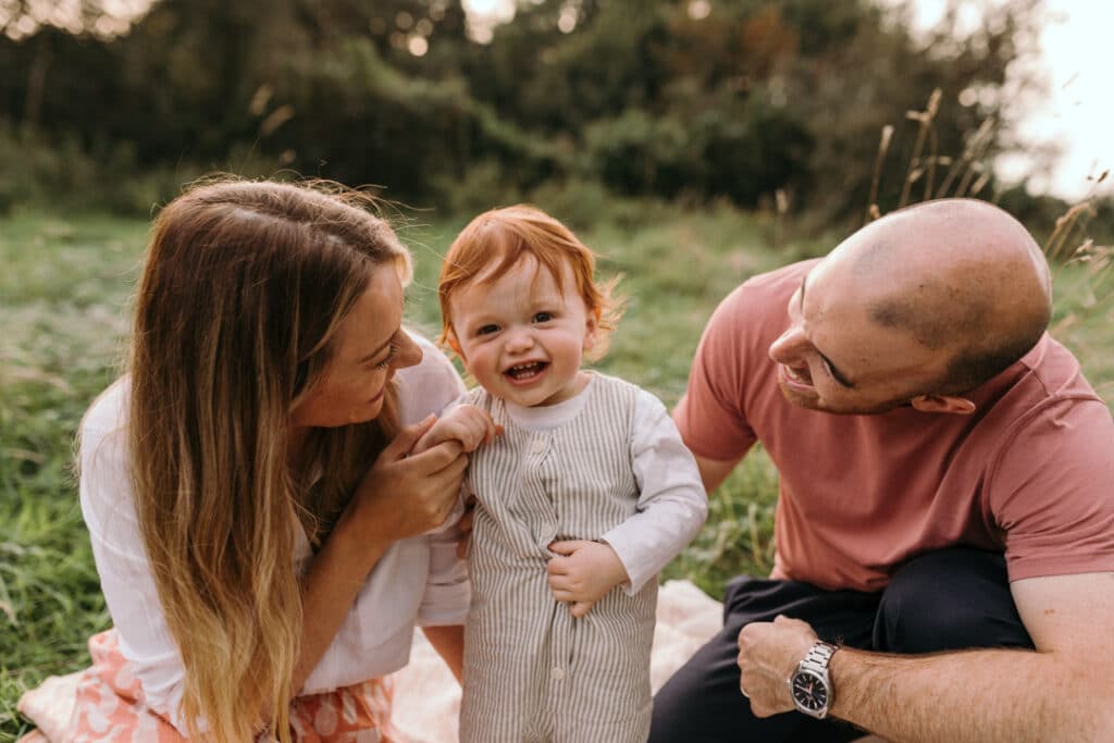 mum and dad are sitting on the blanket and looking at their boy. the boy is looking at the camera and smiling. he is holding his tummy. family photographer in Hampshire. Ewa Jones Photography