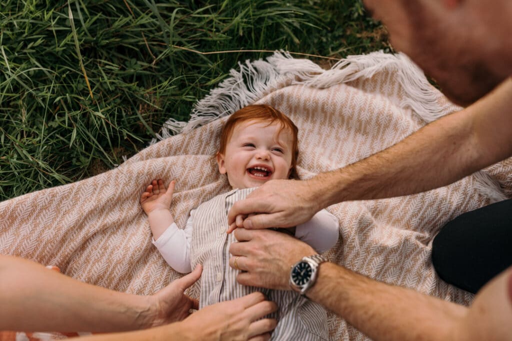 mum and dad are tickling baby boy on his tummy. boy is laying on his back on the blanket and laughing. family photo shoot in Hampshire. Ewa Jones Photography